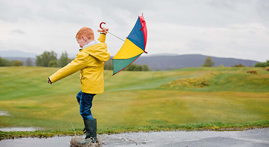 Red-headed boy playing in the rain with an umbrella and rain boots.